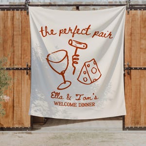 May include: A cream-colored fabric banner featuring rust-colored line art of a wine glass, a corkscrew, and a wedge of cheese. The text reads "the perfect pair" above the illustrations and "Ella & Tom's WELCOME DINNER" printed below them.