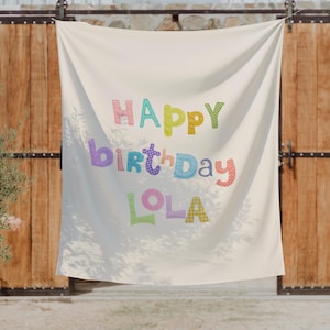 May include: A cream-colored fabric banner featuring the text "HAPPY BIRTHDAY LOLA" in large, multi-colored letters. Each letter displays a unique pattern, including polka dots, checks, and stripes in shades of pink, blue, green, yellow, and orange.