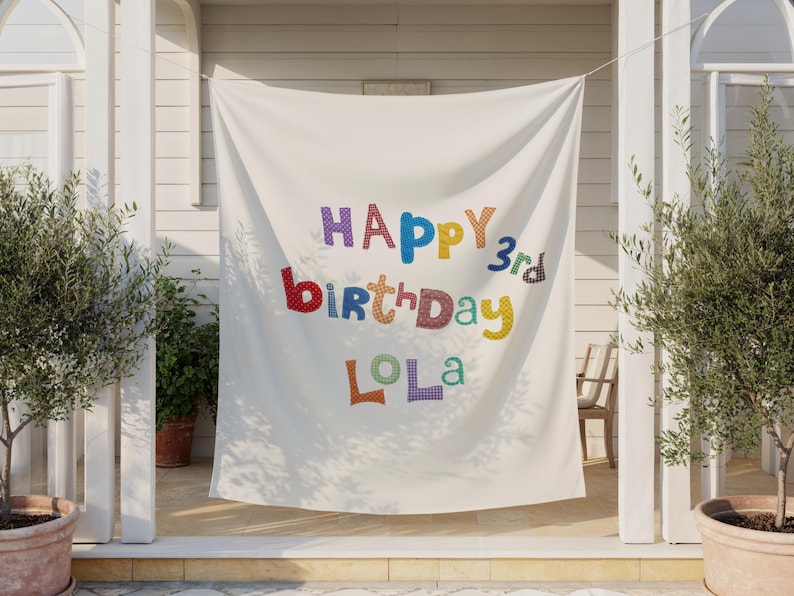 May include: A white fabric banner featuring the colorful text "HAPPY 3rd birthday LOLA" in a playful, multi-patterned font. Each letter displays a unique texture, including polka dots, checkered patterns, and solid colors, hanging as a backdrop.