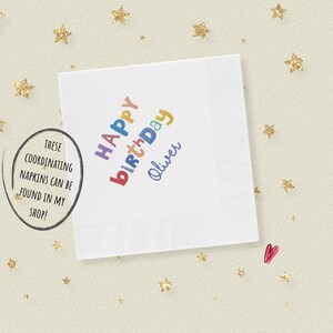 May include: A square white paper napkin featuring the text Happy Birthday Oliver printed in a multi-colored, playful font. The napkin is surrounded by scattered gold glitter stars and a small pink heart icon on a light cream-colored background.