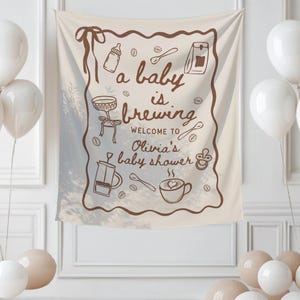 May include: A beige baby shower banner with brown illustrations of coffee-related items and the text "a baby is brewing". White balloons are clustered around the banner, creating a festive atmosphere. The banner is for Olivia's baby shower.