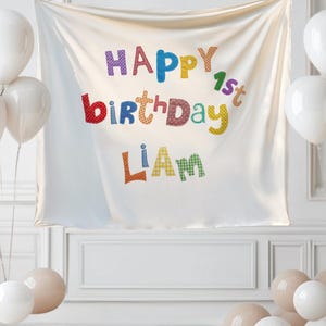 May include: A white fabric backdrop featuring the colorful text Happy 1st Birthday Liam in a playful, patterned font. The banner is surrounded by clusters of white and beige balloons on both sides, creating a festive display for a first birthday.
