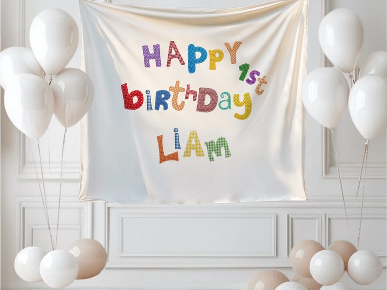 May include: A white fabric backdrop featuring the colorful text Happy 1st Birthday Liam in a playful, patterned font. The banner is surrounded by clusters of white and beige balloons on both sides, creating a festive display for a first birthday.