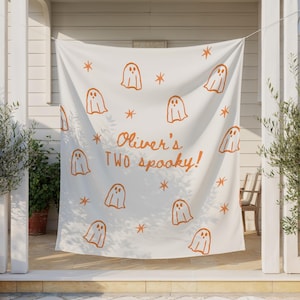 May include: A white fabric backdrop featuring a repeating pattern of orange ghost illustrations and small star shapes. The center of the fabric displays the orange text "Oliver's TWO spooky!" in a handwritten script and block letter style.