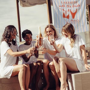 May include: Four women in white t-shirt dresses celebrate with champagne. A banner reads "Lakeside with the Bride" with a bikini graphic. The scene is outdoors, suggesting a bachelorette party or similar event.
