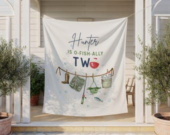 May include: A white fabric backdrop featuring the text Hunter IS O-FISH-ALLY TWO with a red and white fishing bobber replacing the letter O. Below the text, a clothesline displays a brown boot, a green fishing vest, a rod, a hat, and a bucket.
