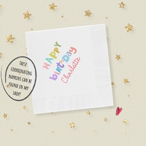 May include: A square white paper napkin featuring the text Happy Birthday Charlotte printed in a multi-colored, playful font. The napkin has a textured border and is surrounded by small gold glitter star shapes and a single small pink heart icon.