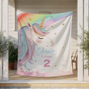 May include: A white fabric backdrop featuring a watercolour unicorn with a gold glitter horn, a pastel rainbow mane, and scattered gold stars. The text reads Eleanor IS TURNING 2 in blue and pink script and block lettering against a soft pink base.