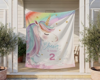 Könnte beinhalten: Ein weißer Stoffhintergrund mit einem Aquarell-Einhorn, einem goldenen Glitzerhorn, einer pastellfarbenen Regenbogenmähne und goldenen Sternen. Der Text lautet Eleanor IS TURNING 2 in blauer und rosa Schrift auf einem hellrosa Untergrund.