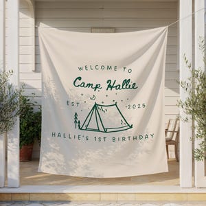 May include: Cream-colored banner with green text and a camping theme. The banner reads "WELCOME TO Camp Hallie EST 2025" with a tent illustration and "HALLIE'S 1ST BIRTHDAY" below. The banner is hanging outdoors.