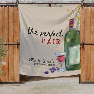 May include: A rectangular fabric banner featuring a painted illustration of a green wine bottle, a glass of red wine, and scattered grapes. The text reads the perfect PAIR and Ella & Tom's REHEARSAL DINNER in black and red script and block lettering.