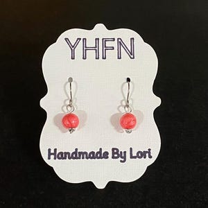 May include: A pair of handmade earrings with red, marbled beads and silver-colored hooks. The earrings are displayed on a white card with the text "YHFH" and "Handmade By Lori" in purple.