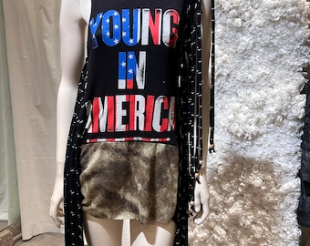 May include: Black sleeveless top with the text "YOUNG IN AMERICA" in red, white, and blue. The top has a faux fur panel at the bottom and long black fringe detailing. The top is displayed on a mannequin.