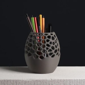 May include: A grey pen holder with a distinctive honeycomb pattern, containing a selection of coloured pens and pencils. The holder is made of a matte material and rests on a white surface against a dark backdrop. Office stationery.