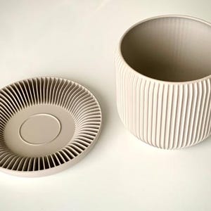 Peut inclure: Un pot de fleurs en céramique beige avec sa soucoupe assortie. Le pot est cylindrique avec des détails côtelés verticaux. La soucoupe présente un motif côtelé en spirale. L'ensemble est sur une surface blanche.