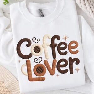 Könnte beinhalten: Weißes Sweatshirt mit gesticktem "Coffee Lover" Design. Das Design zeigt das Wort "Coffee" in Braun-, Beige- und Cremetönen mit Kaffeebohnen-Details. Das Wort "Lover" ist braun. Das Design enthält Sternakzente.