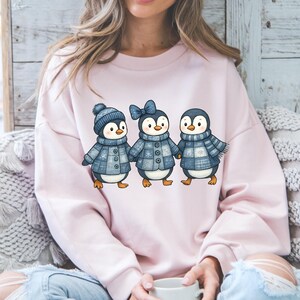 Peut inclure: Un sweat-shirt rose clair avec une illustration à l'aquarelle de trois pingouins. Les pingouins portent des manteaux d'hiver, des chapeaux et des écharpes bleus. Les pingouins se tiennent la main et ont l'air joyeux.