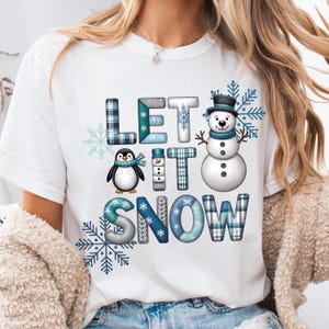 May include: White t-shirt with a winter design. The graphic reads "LET IT SNOW" in different fonts and patterns, accompanied by a penguin, snowman, and snowflakes. The letters and figures use blue and grey plaid and snowflake details.