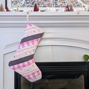May include: A pink and gray Christmas stocking with a white background and a pattern of stripes, zigzags, and gingham. The stocking is hanging on a white fireplace mantel.