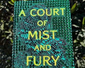May include: A book cover with the title "A Court of Mist and Fury" by Sarah J. Maas. The cover is teal with yellow lettering and a pattern of black dots. The cover is embellished with green rhinestones.