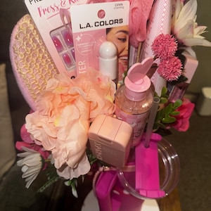 May include: A pink gift basket filled with beauty products and artificial flowers. Items include press-on nails, L.A. Colors products, a loofah, a tumbler, and a small pink purse. The arrangement is set on a white surface.