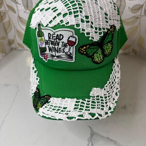 May include: A bright green baseball cap adorned with white crocheted lace trim. The front features an embroidered patch of an open book with a wine bottle and glass, reading READ BETWEEN THE WINES, alongside two green and black butterfly patches.