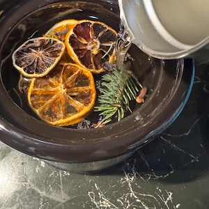 Op de afbeelding: Een donkerbruine slowcooker gevuld met water, gedroogde sinaasappelschijfjes, dennentakjes en kruiden. Er wordt water in de slowcooker gegoten vanuit een zilveren kan. De ingrediënten zijn in de slowcooker gerangschikt.