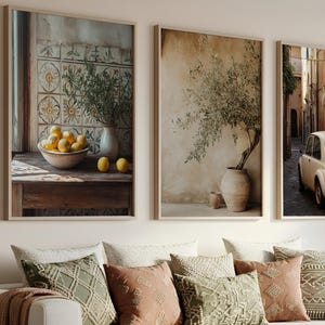 Puede incluir: Tres impresiones artísticas enmarcadas sobre un sofá blanco con cojines decorativos y una manta marrón. Las impresiones representan una naturaleza muerta con limones, un olivo en maceta y una escena callejera con un coche antiguo. Los cojines son en tonos verdes, melocotón y crema.