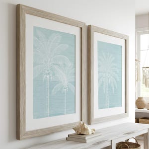 Puede incluir: Dos impresiones artísticas enmarcadas con ilustraciones de palmeras blancas sobre un fondo azul claro. Las impresiones están en marcos de madera clara y se exhiben sobre una consola blanca. Una concha y libros están sobre la mesa.