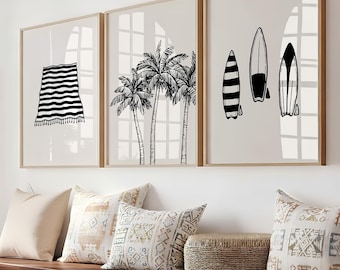 Puede incluir: Tres impresiones artísticas enmarcadas con temática playera. Las impresiones presentan una toalla a rayas blancas y negras, palmeras y tablas de surf. Los marcos son de color marrón claro y el fondo es beige neutro.
