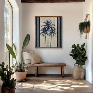 May include: A framed artwork featuring a stylized depiction of three palm trees in navy blue against a cream background. The artwork is framed in a natural wood frame and is displayed above a wooden bench. The room is decorated with plants and natural light.