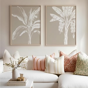 May include: Two framed prints of banana trees in white on a taupe background hang above a white sofa. Cushions in pink, orange, and green with various patterns are arranged on the sofa. A coffee table is in front.