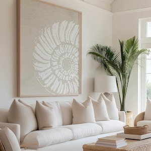 May include: A living room with a large framed artwork featuring a white seashell design on a beige background. A white sofa with several pillows, a wicker coffee table, and a potted palm tree complete the coastal decor.