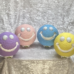 May include: Four colorful smiley face decorative spheres. The spheres are in purple, pink, blue, and yellow. Each sphere has white eyes and a white smiling mouth. The spheres are sitting on a light-colored surface.