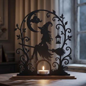 Può includere: Una decorazione a silhouette in metallo nero raffigurante una strega su una scopa, una luna crescente, nuvole e una lanterna sospesa. Il design è incorniciato da intricati viticci e poggia su una base in legno con un lumino acceso.
