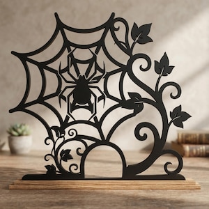 May include: A black laser-cut silhouette of a spider sitting in a web, integrated with swirling vine and leaf patterns. The design is mounted on a rectangular light-colored wooden base, creating a flat, upright decorative display piece.