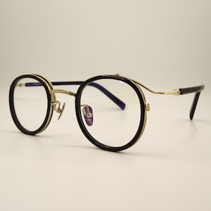 Puede incluir: Gafas redondas con montura negra y detalles dorados. Las lentes son transparentes y las patillas son finas y negras. Las gafas tienen un estilo clásico y vintage.