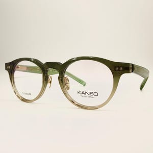 May include: Round-framed eyeglasses in olive green with a gradient tint. The glasses have a titanium frame and the brand name "KANSO TOKYO JAPAN" is printed on the lens. The overall design is modern and stylish.