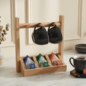May include: Wooden tea station with two black mugs hanging above a tray of tea bags. A cup of tea sits on a coaster, with honey and a spoon nearby. A vase of flowers is on the left.