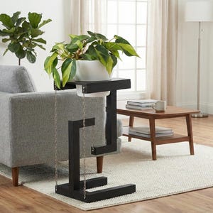 Puede incluir: Soporte de planta negro y moderno con una maceta blanca que contiene una planta verde. El soporte tiene un diseño geométrico único con un detalle de cadena. El soporte se coloca sobre una alfombra blanca en un salón.