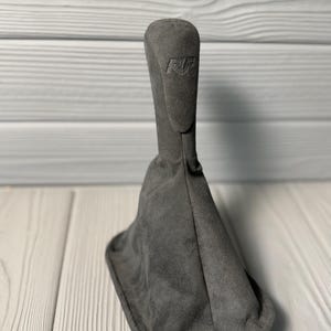 May include: A dark gray gear shift boot with a soft, suede-like texture. The tall, tapered cover features a rectangular base and a vertical design with the letters RF embossed near the top, providing a clean and uniform appearance for vehicle interiors.