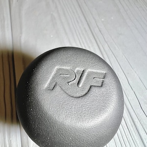 Könnte beinhalten: Ein dunkelgraues, kreisförmiges Objekt mit den Buchstaben "RF" auf der Oberseite. Das Objekt scheint aus einem matten Material zu bestehen und liegt auf einer hellgrauen Holzoberfläche.