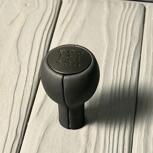 May include: A black gear shift knob with a stitched leather appearance. The top of the knob has the gear pattern 1-3-5-R-2-4. The knob is designed for a manual transmission.
