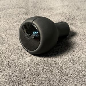 May include: A black shift knob with a shiny, chrome-like center. The knob is round with a cylindrical base and is made of a smooth, possibly leather-like material. The center has the letters "RU" engraved.