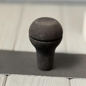 May include: A dark gray, textured shift knob with a rounded top and a cylindrical base. The knob has a slightly rough surface and a seam around the middle. The background is blurred, with a light-colored wooden surface.
