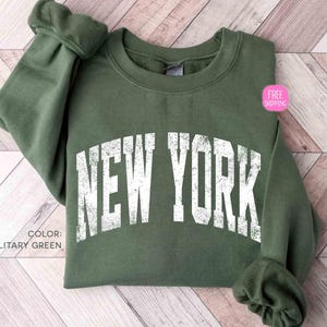 Può includere: Felpa verde militare con la scritta "NEW YORK" in lettere bianche consumate. La maglia a girocollo a maniche lunghe è esposta su una superficie di legno chiaro. È visibile un adesivo rosa con la scritta "SPEDIZIONE GRATUITA".