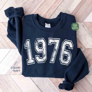 Può includere: Felpa blu navy con l'anno "1976" stampato in bianco, con un effetto invecchiato. La felpa ha un girocollo e maniche lunghe con polsini a coste. Il colore è blu navy.