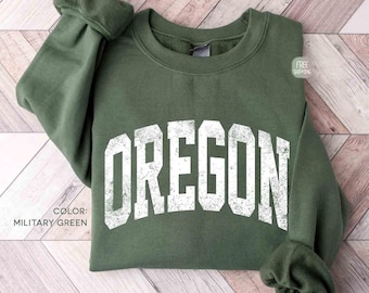 May include: A military green crewneck sweatshirt featuring the word OREGON printed in a large, distressed white collegiate font across the chest. The long-sleeved garment has ribbed cuffs and a ribbed neckline, showcasing a simple, solid design.