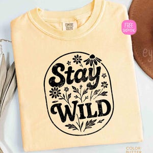 May include: A light yellow t-shirt with a crew neck. The shirt features a black oval design with the words "Stay Wild" and floral accents. The shirt is labeled "Comfort Colors" and the color is "Butter".