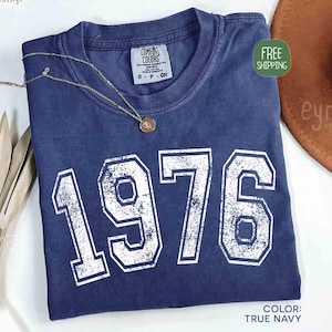 May include: A navy blue t-shirt with the year "1976" printed in white, featuring a crew neck and short sleeves. A gold necklace with a pendant rests on the shirt. The shirt is labeled "Comfort Colors" and the color is "True Navy".